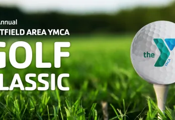 A close-up of a golf ball with the YMCA logo resting on a tee in bright green grass, with text on the left reading "21st Annual Westfield Area YMCA Golf Classic."