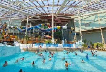 Indoor wave pool at Aquatopia Waterpark with swimmers in the water, colorful waterslides in the background, and a large glass roof overhead.
