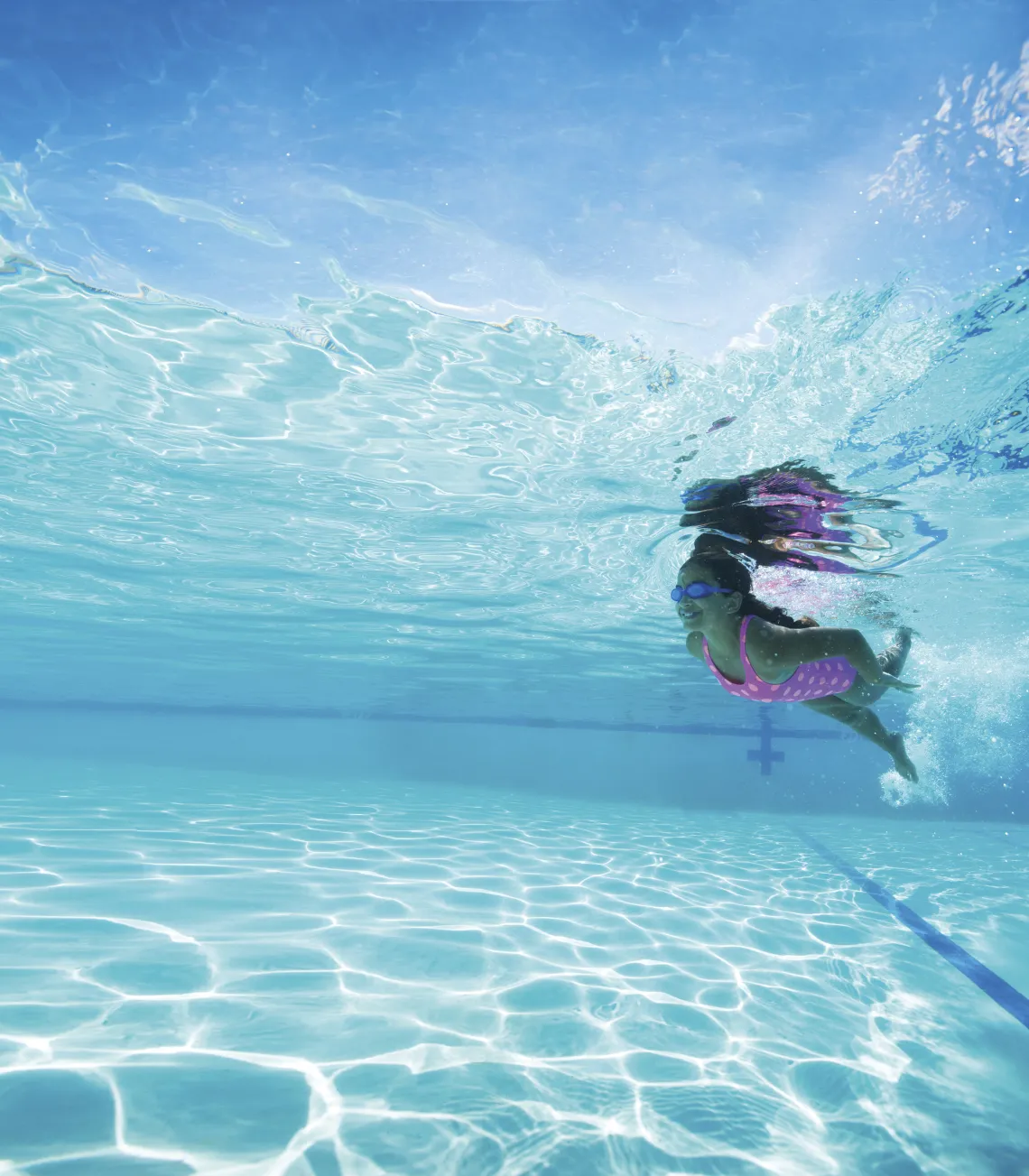 Child swimming underwater in a pool during swim practice, wearing goggles and a pink swimsuit.