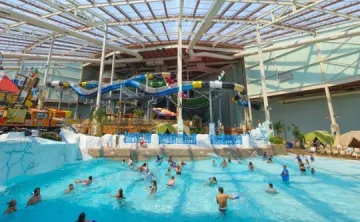 Indoor wave pool at Aquatopia Waterpark with swimmers in the water, colorful waterslides in the background, and a large glass roof overhead.